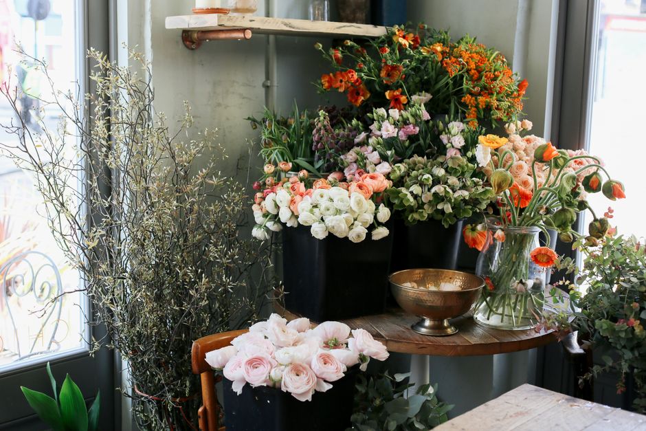 A collection of fresh floral arrangements on a wooden table near a window, featuring a variety of flowers in shades of white, pink, orange, and purple. The arrangement includes pale pink roses and whi
